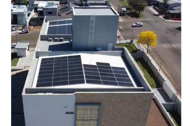 Foto horizontal: prédio com painéis solares no telhado, área urbana ao redor, destaque para energia limpa e sustentabilidade.