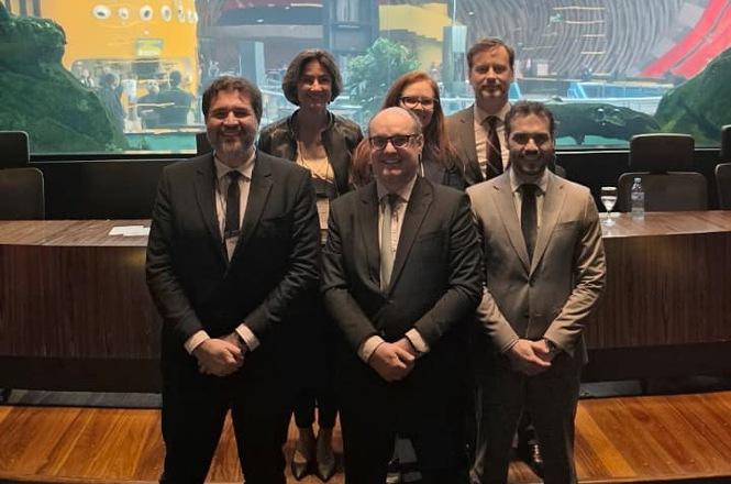 Foto horizontal: seis autoridades posam diante de mesa em auditório, com painel colorido ao fundo.
