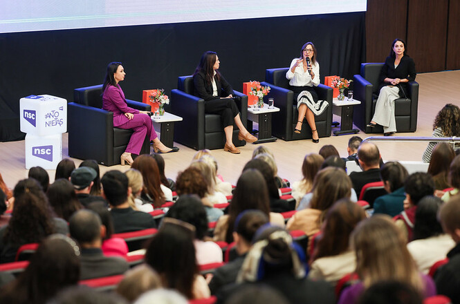 A imagem mostra um painel de quatro mulheres em poltronas, discutindo em um palco com flores e logotipos ao lado, enquanto a plateia atenta acompanha no auditório.