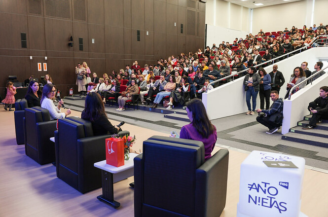 A imagem mostra um auditório lotado, com quatro mulheres em poltronas no palco participando de um painel, enquanto a plateia, em cadeiras vermelhas, acompanha atenta em clima de concentração e aprendizado.