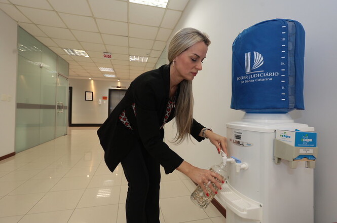  Copilot said: Horizontal: Pessoa enche garrafa reutilizável em bebedouro instalado em corredor institucional, com piso cerâmico claro, paredes brancas, divisórias de vidro, iluminação embutida no teto e elevadores ao fundo. Ao lado do equipamento, há dispensador de copos reutilizáveis identificado com a marca do Poder Judiciário de Santa Catarina.