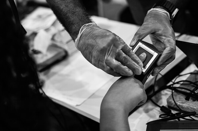 Foto Preto e Branco: mãos em leitor biométrico sobre mesa, ambiente interno, atendimento público, coletando digitais em preto e branco