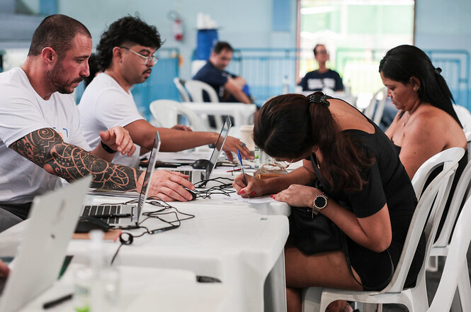 Foto colorida de atendimento com pessoas, salão interno azul e branco, ação social, preenchendo cadastro