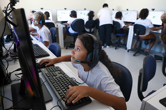 Criança usa computador com fone em sala de informática, concentrada em atividade digital e aprendizado tecnológico