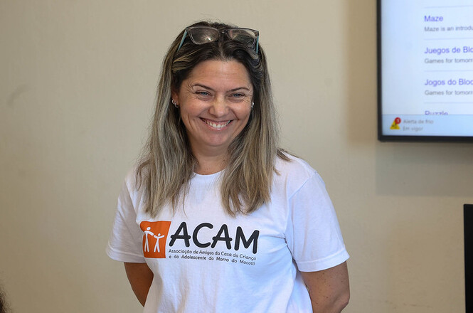 Mulher sorri em ambiente educacional, camiseta da ACAM em destaque, clima de acolhimento e atuação social