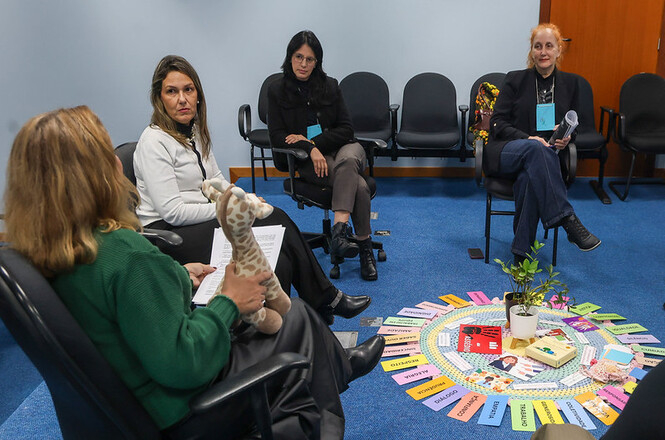 Foto horizontal: mulheres participam de dinâmica em círculo, uma facilitadora segura objeto durante fala, materiais ao centro, ambiente interno, clima de escuta e reflexão.
