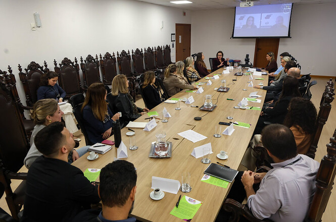 Foto horizontal: reunião institucional com participantes ao redor de mesa, apresentação projetada ao fundo, equipe atenta à discussão, ambiente formal, clima de diálogo e alinhamento.