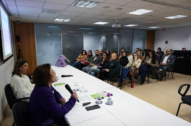 Foto horizontal: mesa de debate com participantes em evento institucional, mediadora fala ao microfone enquanto público acompanha atento, ambiente formal, clima de escuta e reflexão coletiva.