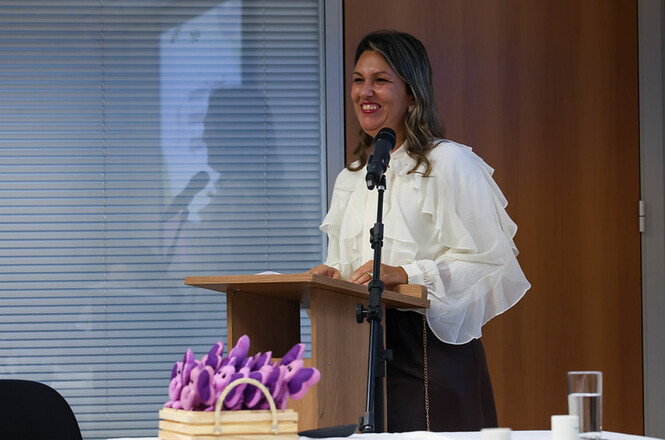 Foto horizontal: mulher discursa ao microfone em evento institucional, expressão sorridente ao púlpito, público ao redor, ambiente formal, clima de acolhimento e diálogo.