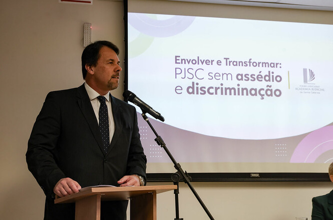 Foto horizontal: autoridade discursa ao microfone durante evento institucional do PJSC, apresentação sobre combate ao assédio e discriminação projetada ao fundo, ambiente formal, clima de conscientização e compromisso.