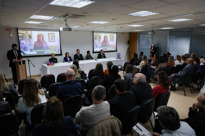 Foto horizontal: público acompanha mesa de debate em evento institucional, autoridades à frente e transmissão exibida em telões, ambiente formal, clima de reflexão e diálogo coletivo.
