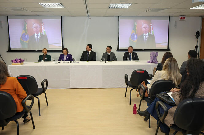 Foto horizontal: mesa de autoridades participa de evento institucional, público acompanha ao fundo e transmissão exibida em telões, ambiente formal, clima de reflexão e diálogo coletivo.