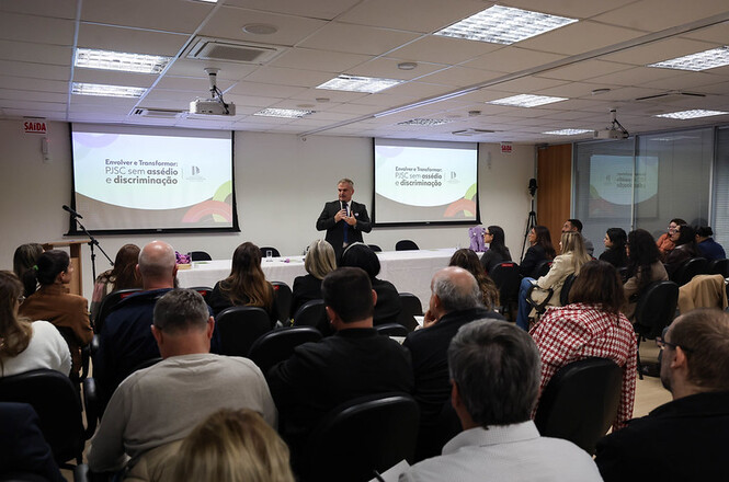 Foto horizontal: público acompanha palestra em evento institucional do PJSC, expositor ao centro e apresentação sobre combate ao assédio e discriminação projetada em telões, ambiente formal, clima de conscientização