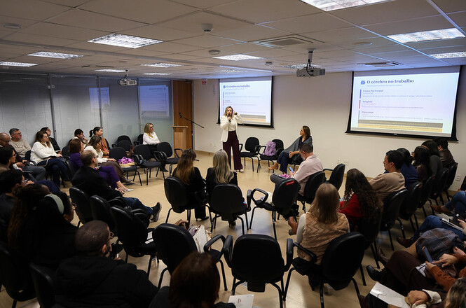 Foto horizontal: participantes em dinâmica de grupo durante capacitação, facilitadora conduz atividade ao centro com apoio de projeções, pessoas em círculo acompanham, ambiente institucional