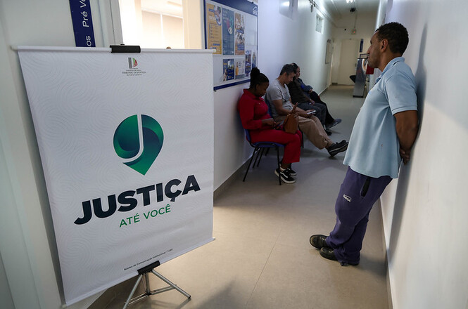 Foto horizontal: pessoas aguardam atendimento em corredor com banner “Justiça Até Você”, clima de orientação.
