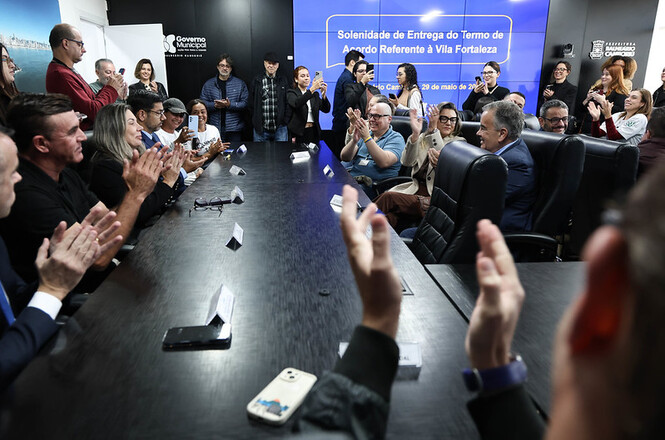 Foto horizontal: participantes aplaudem durante solenidade de entrega em sala institucional, clima de celebração e reconhecimento coletivo.