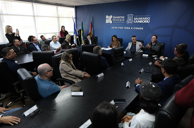 Foto horizontal: autoridades e representantes participam de reunião em mesa ampliada na Prefeitura de Balneário Camboriú, debate coletivo em andamento, clima de articulação e cooperação institucional.