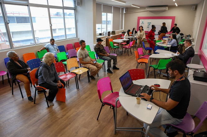 Foto horizontal: atendimento em espaço comunitário com pessoas aguardando e equipe prestando orientação.