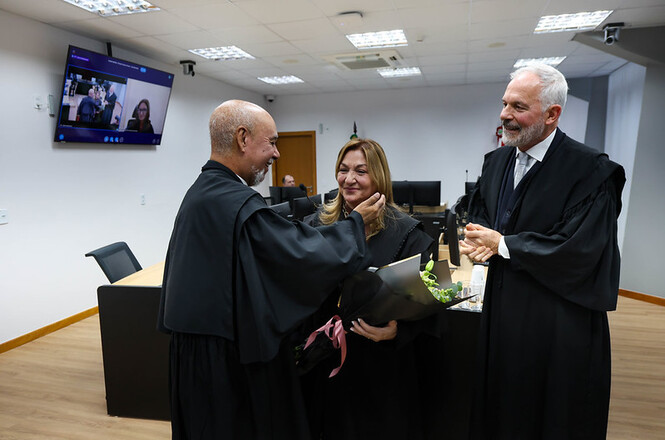 Foto horizontal: magistrados participam de homenagem em ambiente judicial, gesto afetuoso e entrega de flores em destaque, clima de reconhecimento, despedida e valorização da trajetória profissional.