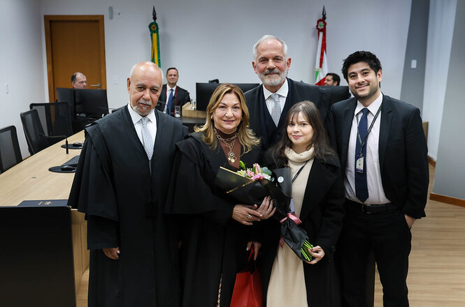 Foto horizontal: magistrados e familiares posam em ambiente judicial durante homenagem de aposentadoria, buquê de flores em destaque, clima de reconhecimento, celebração e encerramento de ciclo profissional.