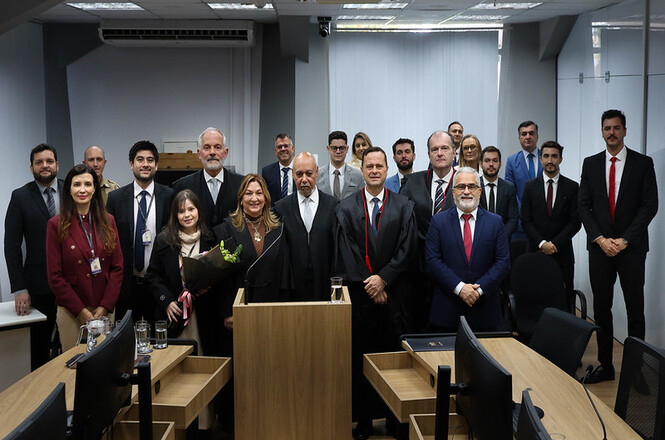 Foto horizontal: grupo de magistrados, servidores e familiares reunidos em ambiente judicial durante homenagem de aposentadoria, destaque para a homenageada ao centro, clima de reconhecimento, celebração coletiva e encerramento de trajetória profissional.