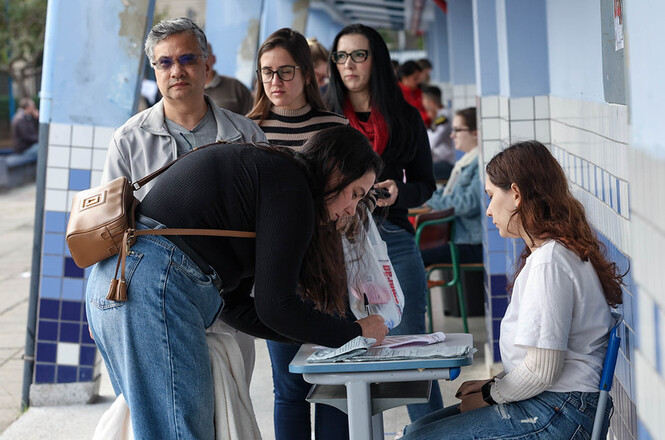 Foto horizontal: candidata assina lista de presença enquanto fiscais organizam atendimento a participantes em corredor escolar.