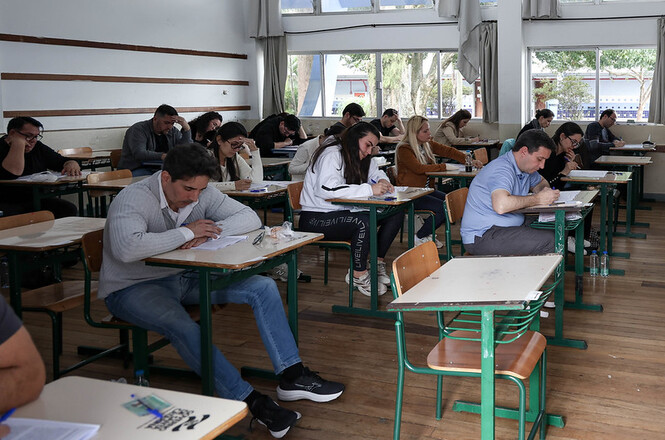 Foto horizontal: candidatos realizam prova em sala de aula, concentrados em carteiras distribuídas pelo ambiente.