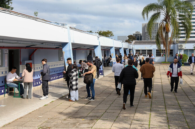 Foto horizontal: candidatos circulam e aguardam atendimento em área externa de local de prova, entre salas de aplicação.