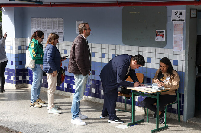 Foto horizontal: candidato assina lista de presença enquanto fiscais atendem participantes em corredor escolar.