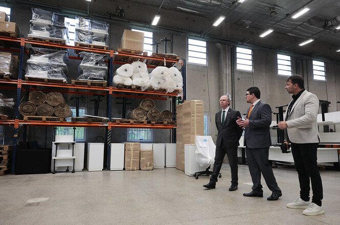 Foto horizontal: pessoas em tons preto, cinza e branco em galpão industrial, ambiente corporativo, realizando visita técnica entre estantes e equipamentos.