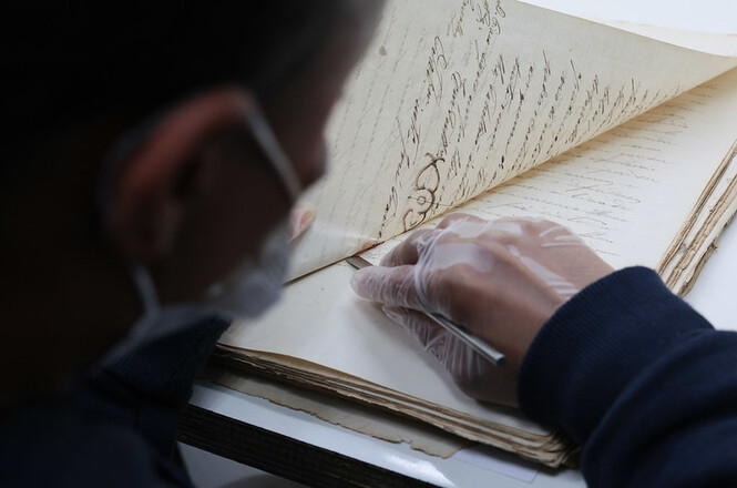 Foto horizontal: profissional utiliza luvas e máscara para manusear documento histórico durante trabalho de preservação do acervo documental.