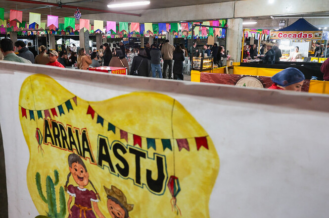 Foto horizontal: cartaz do "Arraiá ASTJ" em primeiro plano e público circulando entre barracas durante a festa junina promovida pelo TJSC.