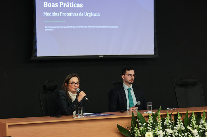 Foto horizontal: dois palestrantes participam de painel sobre boas práticas na concessão de medidas protetivas de urgência, durante evento institucional voltado ao enfrentamento da violência doméstica contra a mulher.