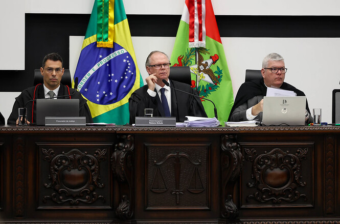 Foto horizontal: o presidente do Tribunal de Justiça de Santa Catarina, desembargador Rubens Schulz, conduz sessão do Tribunal Pleno ao lado do procurador de Justiça e do secretário da sessão, com as bandeiras do Brasil e de Santa Catarina ao fundo.