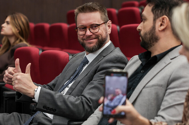 Imagem horizontal mostra magistrado sentado em auditório, sorrindo e conversando com homem ao lado.