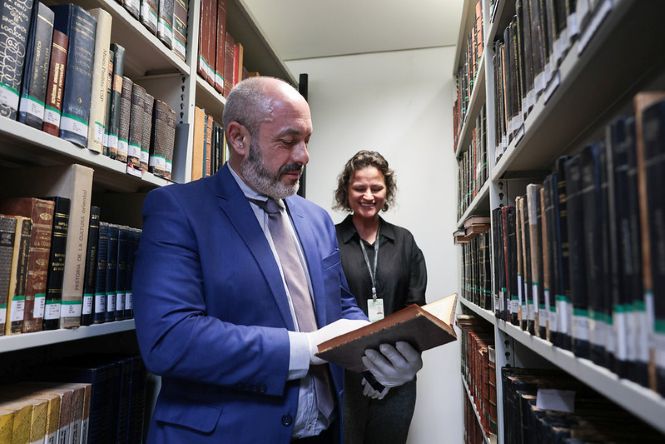 Foto horizontal: 1º vice-presidente do TJSC, desembargador André Luiz Dacol, de terno azul e luvas brancas, consulta obra rara entre estantes de livros da biblioteca.