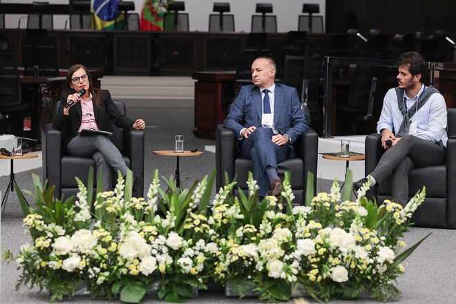 Foto horizontal: autoridade fala ao microfone durante debate no plenário, acompanhada por outros dois participantes.