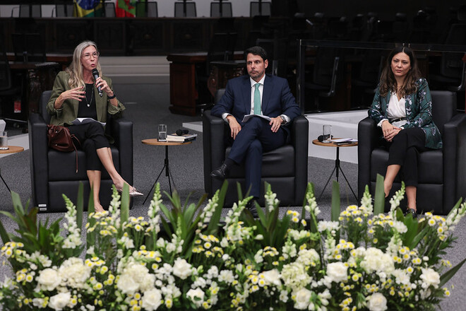 Foto horizontal: autoridade de blazer verde fala ao microfone; ao lado, homem de terno azul e mulher de blazer estampado.**