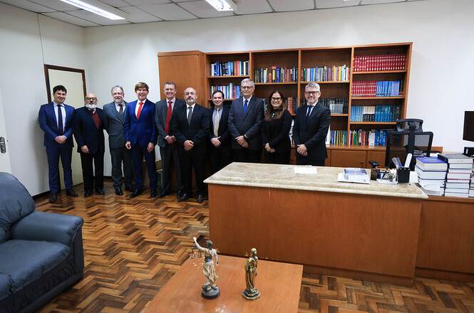 Foto horizontal: autoridades posam em escritório, diante de estante com livros; mesa exibe esculturas e documentos.