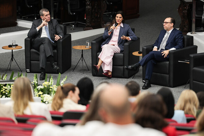 Foto horizontal: autoridade de blazer azul e vestido rosa fala ao microfone; dois homens de ternos escuros acompanham.