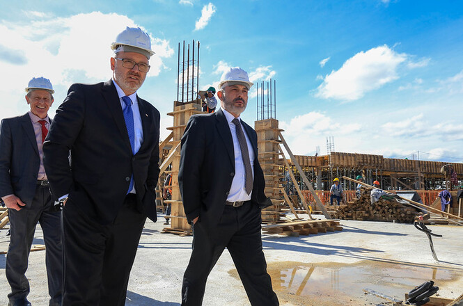 Foto horizontal: des. André Luiz Dacol, de terno preto, gravata marrom e capacete branco, caminha pelo canteiro de obras.