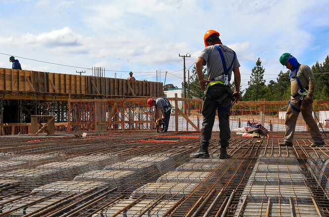 Foto horizontal: trabalhadores com capacetes e equipamentos de segurança atuam sobre estrutura metálica em canteiro de obras.