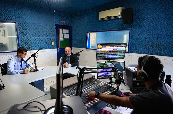 Foto horizontal: estúdio de transmissão reúne microfone, mesas de áudio, monitores e equipamentos eletrônicos.