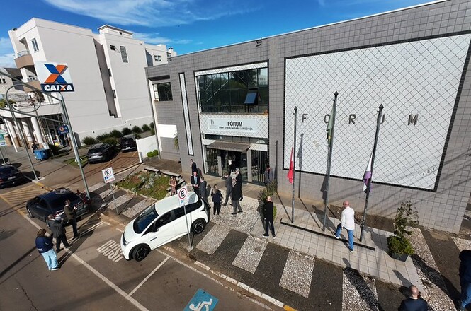 Foto horizontal: fachada de fórum, com bandeiras, veículos e pessoas na entrada; agência da Caixa aparece ao lado.