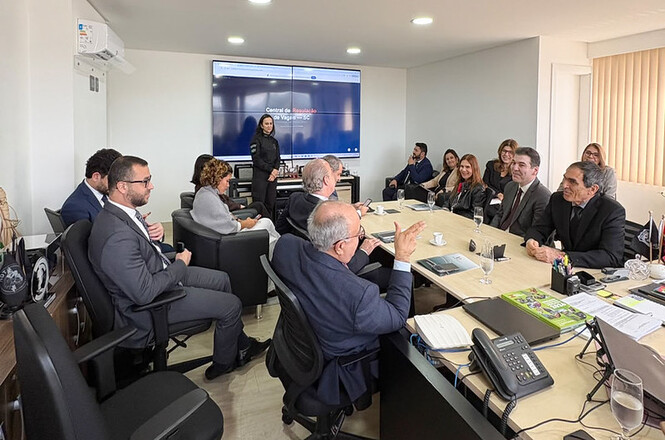 Foto horizontal: autoridades discutem durante reunião, com policial ao fundo e tela sobre a Central de Regulação de Vagas.