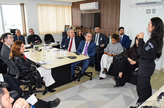 Foto horizontal: policial de uniforme preto faz exposição a autoridades reunidas em sala, com documentos e copos à mesa.