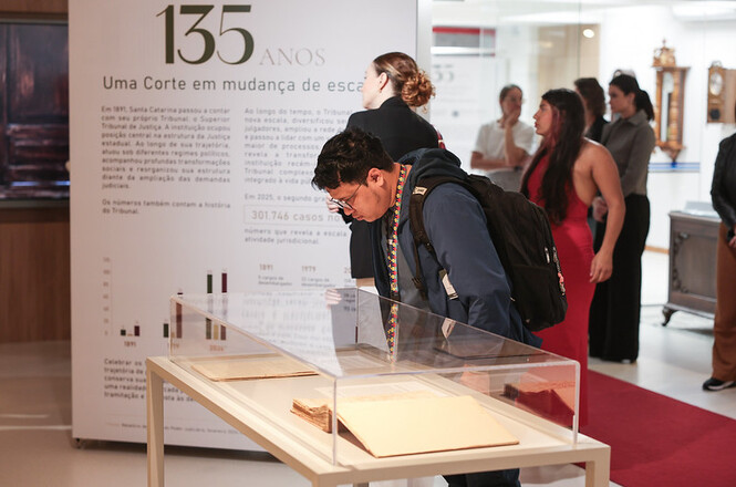 Foto horizontal: visitante observa documentos históricos expostos em vitrine durante mostra comemorativa dos 135 anos do TJSC.