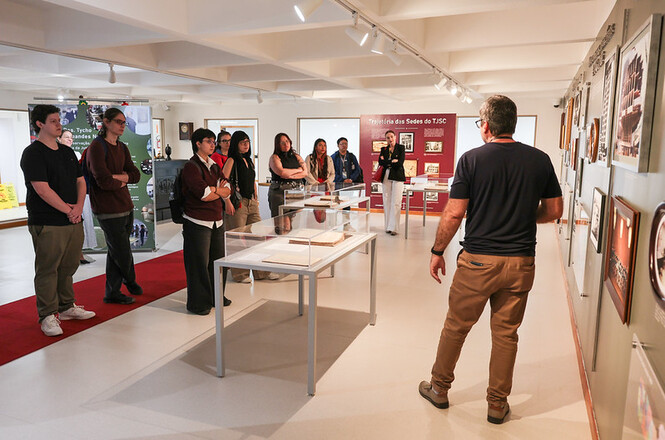 Foto horizontal: visitantes acompanham visita guiada em exposição sobre a trajetória histórica das sedes do TJSC.