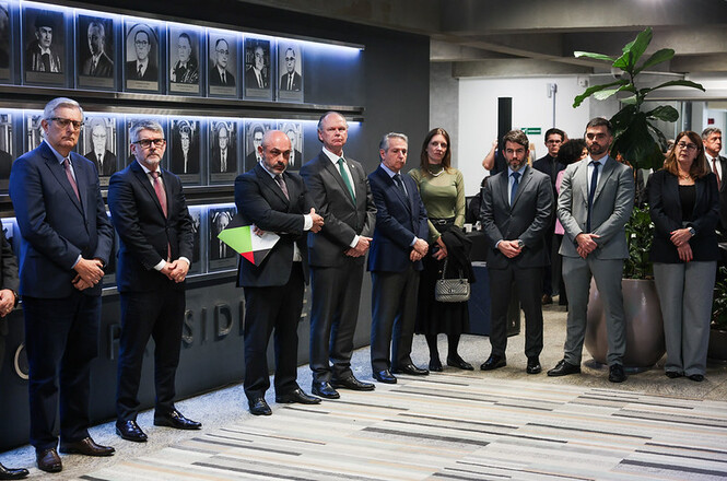 Foto horizontal: segurando uma pasta verde, o desembargador André Luiz Dacol posa ao lado do presidente do TJSC, desembargador Rubens Schulz, e de outras autoridades, diante da galeria de retratos de ex-presidentes do Tribunal.