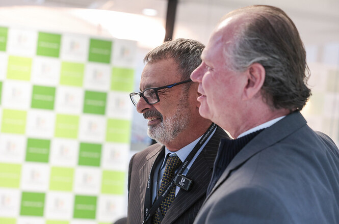 Foto horizontal: oficial de justiça Paulo Roberto Costa, de terno marrom, gravata e óculos, aparece sorrindo ao lado do presidente do TJSC, desembargador Rubens Schulz, ambos de perfil, diante de painel institucional verde e branco do Poder Judiciário de Santa Catarina.